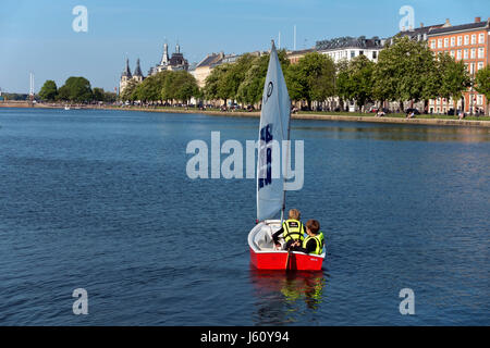 Due ragazzi in barca a vela un ottimista in gommone sul lago Peblinge nel centro di Copenhagen in un assolato pomeriggio di inizio dell'estate. La gente a prendere il sole sulla banca. Foto Stock