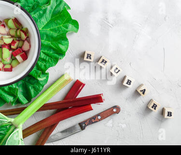 Fresche biologiche degli steli di rabarbaro con pezzi tagliati nel recipiente di metallo al di sopra di cemento grigio sfondo di pietra. Parola di rabarbaro scritto con blocchi di legno lettere Foto Stock