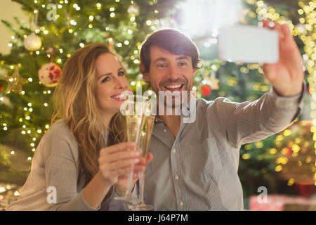 Coppia sorridente tostare champagne flauti e tenendo selfie davanti dell albero di Natale Foto Stock