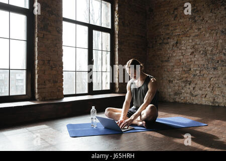 Immagine di bello forte atleta seduto vicino a bottiglia di acqua sul pavimento in palestra utilizzando computer portatili. Guardando a parte. Foto Stock