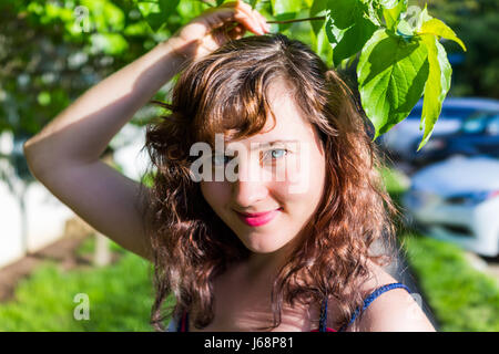 Closeup Ritratto di giovane brunette volto di donna con colore rosso o rosa rossetto sorridente sotto verde foglie in estate Foto Stock