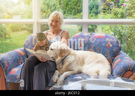 Bionda sorridente donna di mezza età è la lettura di un libro seduti sul divano insieme con un gatto e il Golden Retriever cane. Gli animali stanno guardando il libro. Foto Stock