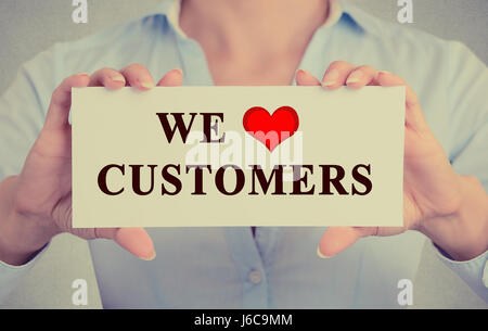 Primo piano immagine retrò imprenditrice mani segno bianco o una scheda con il messaggio che amiamo i clienti isolate su ufficio grigio Sfondo a parete Foto Stock