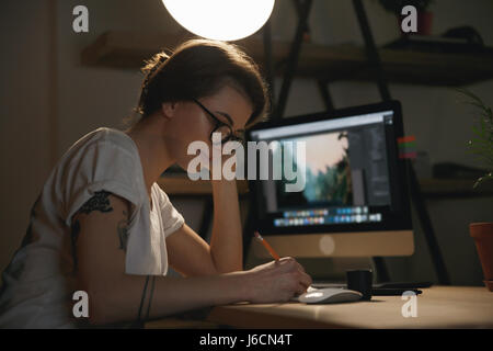 Immagine della signora giovane designer seduto al chiuso di notte schizzi di disegno in un album mentre si utilizza il computer. Foto Stock