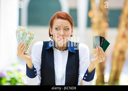 Closeup ritratto confuso business donna con carte di credito in una mano e in contanti, le fatture del dollaro in altri, perplesso che cosa è meglio città isolata backgrou Foto Stock