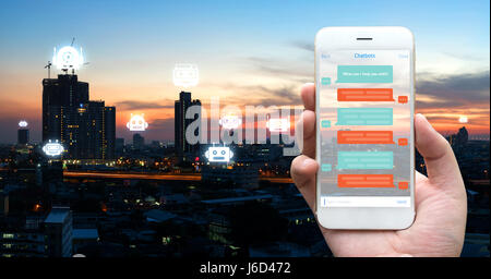Chat bot , Robo ovunque e futuro concetto di commercializzazione. Mano umana azienda telefono bianco , l'interfaccia utente delle applicazioni e icone di robot con la città sullo sfondo del tramonto Foto Stock