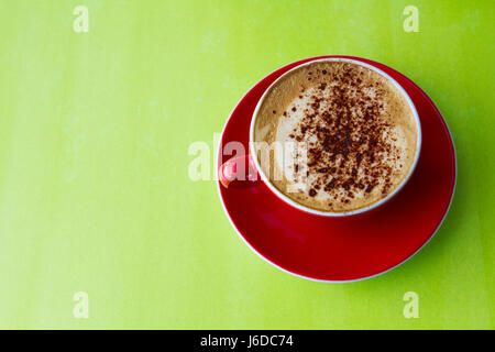 Guardando verso il basso a partire da sopra su una schiumosa tazza di cappuccino caffè in un bicchiere di rosso e il piattino e su un colorato, superficie verde. Foto Stock