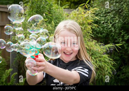 Dieci anni di ragazza bionda utilizza una macchina piccola per soffiare le bolle in corrispondenza della telecamera in un giardino Foto Stock