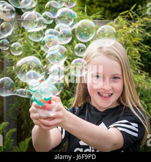 Dieci anni di ragazza bionda utilizza una macchina piccola per soffiare le bolle in corrispondenza della telecamera in un giardino Foto Stock