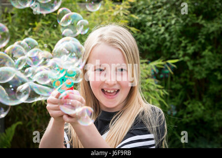 Dieci anni di ragazza bionda utilizza una macchina piccola per soffiare le bolle in corrispondenza della telecamera in un giardino Foto Stock
