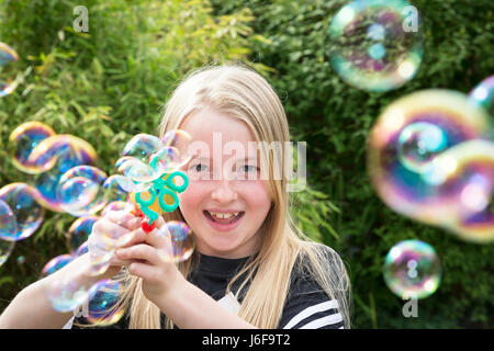 Dieci anni di ragazza bionda utilizza una macchina piccola per soffiare le bolle in corrispondenza della telecamera in un giardino Foto Stock