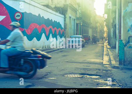 Immagine granulosa back street motociclista passa fuori del telaio in movimento sfocato mentre blu classico americano auto parcheggiate e carretti a mano può essere visto ulteriori Foto Stock