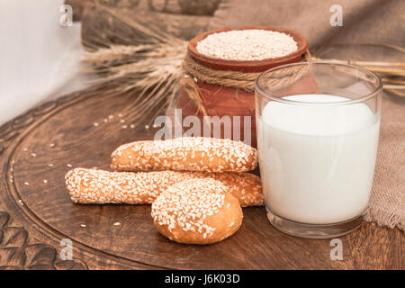 Latte in vetro, grissini, panini su un tavolo di legno. Latte di sesamo in vetro. White Semi di sesamo nella pentola di creta su un tavolo di legno. Una sana colazione e bever Foto Stock