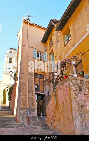 La vecchia strada collinare a Villefranche-sur-mer con case inclinato e la torre dell'orologio di st michel chiesa sullo sfondo, Francia. Foto Stock