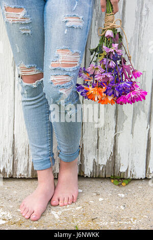 A piedi nudi ragazza adolescente in Sfrangiati Jeans blu con colorati daisy bouquet da fienile in legno Foto Stock