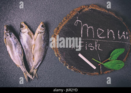 Tre pesci secchi su una superficie nera, accanto ad un moncone di legno con una scritta del menu Chalk Foto Stock