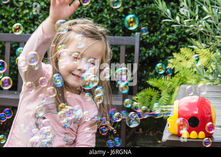 Bimba di tre anni giocando a bolle costituito da una macchina di bolle in un giardino. Sussex, Regno Unito. Foto Stock