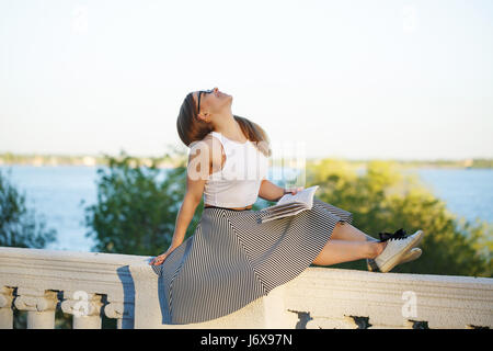 Giovane ragazza attraente hipster è la lettura di un libro. Ella è vestita in bicchieri, top, gonna e scarpe da ginnastica. Una studentessa è impegnato in auto-educazione. Studio Foto Stock