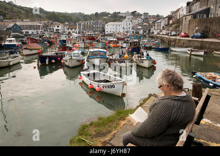 Regno Unito, Cornwall, Mevagissey, donna visitatore seduto sul banco harbourside libro di lettura nella luce del sole Foto Stock
