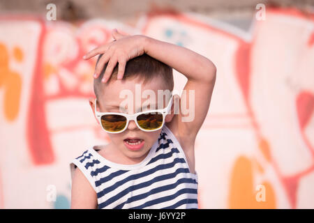 Adorable little boy in occhiali da sole e sailor stripes shirt su giallo e rosa graffiti sullo sfondo della parete tenendo la sua testa con la mano Foto Stock