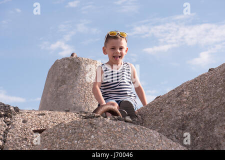 Ragazzino con gli occhiali da sole e sailor stripes shirt udienza del frangiflutti e che mostra la linguetta su sfondo cielo Foto Stock