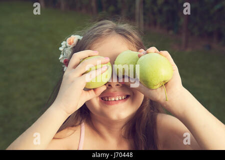 Una giovane ragazza allegra tiene giocosamente mele verdi sugli occhi, Foto Stock