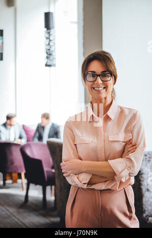 Business donna in piedi con le braccia piegate. Lei è sorridente alla fotocamera. Due uomini in giacca può essere visto a parlare in background. Foto Stock