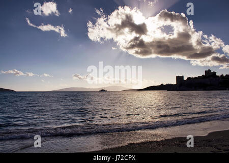 Sun luce irrompe attraverso le nuvole in cielo drammatico. Essa brilla sulla superficie del mare a Bodrum Turchia sudoccidentale. La luce di retromarcia rendono il castello di Bodrum guarda come silh Foto Stock