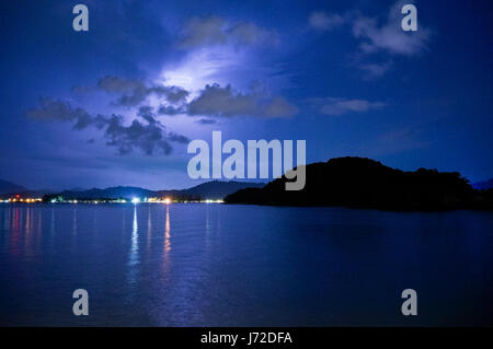 Un fulmine su un mare calmo, nuvole scure e un'isola. Luci lontane formano una città sono visibili anche in questo night shot Foto Stock