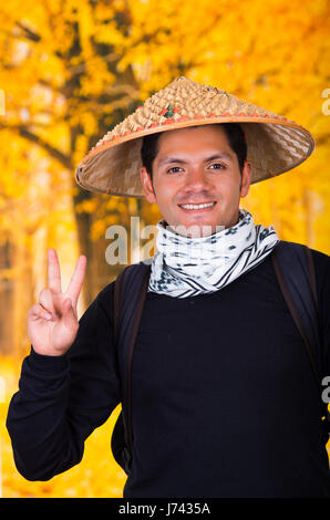 Ritratto di un bel giovane ispanica business ragazzo indossa un asiatico cappello conico e facendo un segnale di vittoria nella collezione autunno sfondo Foto Stock