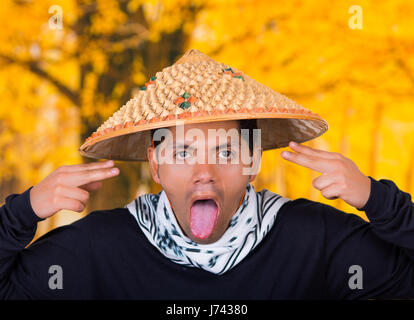 Close up di un bel giovane ispanica business ragazzo indossa un asiatico cappello conico facendo una buffa faccia ed estrarre la linguetta nella collezione autunno sfondo Foto Stock