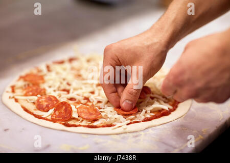 Cuocere le mani aggiungendo salami di pizza in pizzeria Foto Stock