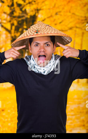 Ritratto di un bel giovane ispanica business ragazzo indossa un asiatico cappello conico facendo una buffa faccia ed estrarre la linguetta nella collezione autunno sfondo Foto Stock