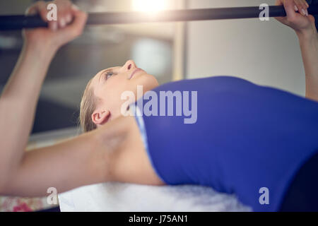 Close-up vista laterale della giovane donna bionda in blu in alto facendo panca esercizio di pressatura con bar-campana in palestra Foto Stock