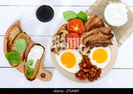 Prima colazione inglese: uova, pancetta, fagioli in salsa di pomodoro, funghi, pomodori, toast con crema di formaggio e una tazza di caffè su un bianco sullo sfondo di legno. Foto Stock