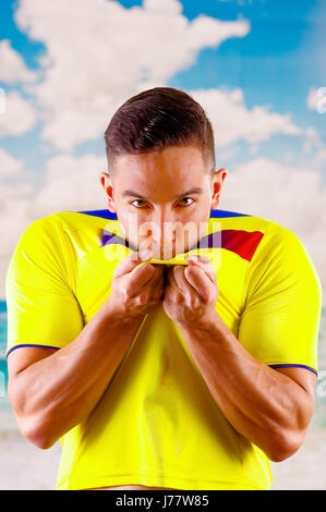 Giovane uomo ecuadoriana indossando maratona ufficiale Football Shirt in piedi di fronte alla fotocamera, molto impegnato il linguaggio del corpo la visione di gioco con grande entusiasmo, cielo blu e nuvole sullo sfondo Foto Stock