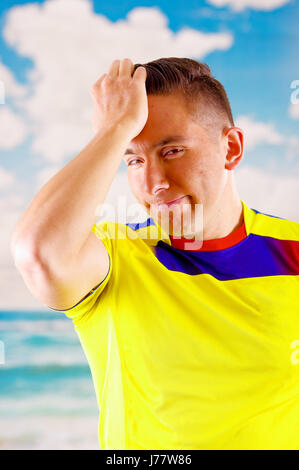 Giovane uomo ecuadoriana indossando maratona ufficiale Football Shirt in piedi di fronte alla fotocamera, molto impegnato il linguaggio del corpo la visione di gioco con grande entusiasmo, cielo blu e nuvole sullo sfondo Foto Stock