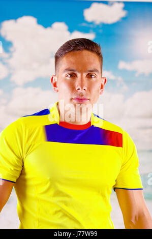 Giovane uomo ecuadoriana indossando maratona ufficiale Football Shirt in piedi di fronte alla fotocamera, molto impegnato il linguaggio del corpo la visione di gioco con grande entusiasmo, cielo blu e nuvole sullo sfondo Foto Stock