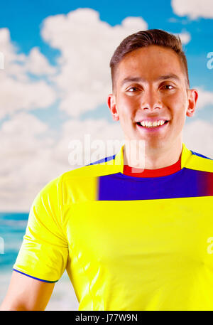 Giovane uomo ecuadoriana indossando maratona ufficiale Football Shirt in piedi di fronte alla fotocamera, molto impegnato il linguaggio del corpo la visione di gioco con grande entusiasmo, cielo blu e nuvole sullo sfondo Foto Stock