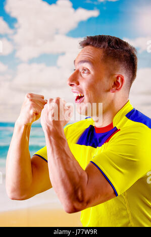 Giovane uomo ecuadoriana indossando maratona ufficiale Football Shirt in piedi di fronte alla fotocamera, molto impegnato il linguaggio del corpo la visione di gioco con grande entusiasmo, cielo blu e nuvole sullo sfondo Foto Stock