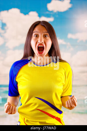 Giovane donna ecuadoriana indossando maratona ufficiale Football Shirt in piedi di fronte alla fotocamera, molto impegnato il linguaggio del corpo la visione di gioco con grande entusiasmo, cielo blu e nuvole sullo sfondo Foto Stock