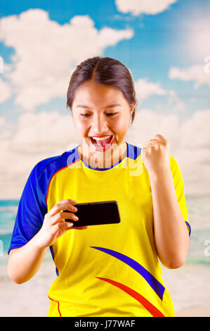 Giovane donna ecuadoriana indossando maratona ufficiale Football Shirt in piedi di fronte alla fotocamera, molto impegnato il linguaggio del corpo la visione di gioco con grande entusiasmo, cielo blu e nuvole sullo sfondo Foto Stock