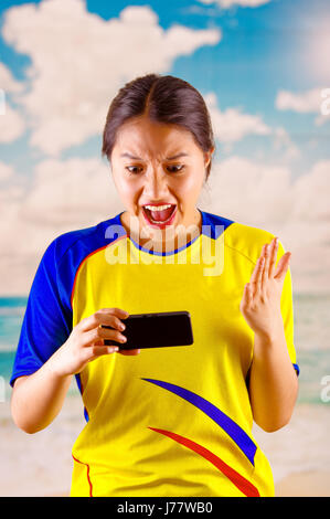 Giovane donna ecuadoriana indossando maratona ufficiale Football Shirt in piedi di fronte alla fotocamera, molto impegnato il linguaggio del corpo la visione di gioco con grande entusiasmo, cielo blu e nuvole sullo sfondo Foto Stock