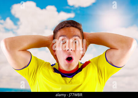 Giovane uomo ecuadoriana indossando maratona ufficiale Football Shirt in piedi di fronte alla fotocamera, molto impegnato il linguaggio del corpo la visione di gioco con grande entusiasmo, cielo blu e nuvole sullo sfondo Foto Stock