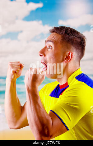 Giovane uomo ecuadoriana indossando maratona ufficiale Football Shirt in piedi di fronte alla fotocamera, molto impegnato il linguaggio del corpo la visione di gioco con grande entusiasmo, cielo blu e nuvole sullo sfondo Foto Stock