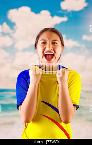 Giovane donna ecuadoriana indossando maratona ufficiale Football Shirt in piedi di fronte alla fotocamera, molto impegnato il linguaggio del corpo la visione di gioco con grande entusiasmo, cielo blu e nuvole sullo sfondo Foto Stock