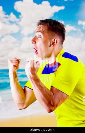 Giovane uomo ecuadoriana indossando maratona ufficiale Football Shirt in piedi di fronte alla fotocamera, molto impegnato il linguaggio del corpo la visione di gioco con grande entusiasmo, cielo blu e nuvole sullo sfondo Foto Stock