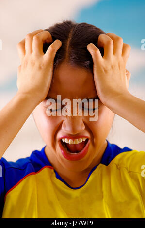 Giovane donna ecuadoriana indossando maratona ufficiale Football Shirt in piedi di fronte alla fotocamera, molto impegnato il linguaggio del corpo la visione di gioco con grande entusiasmo, cielo blu e nuvole sullo sfondo Foto Stock