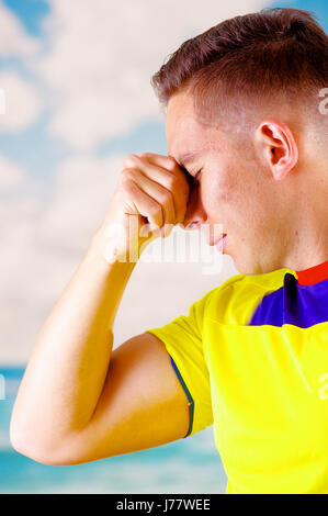 Giovane uomo ecuadoriana indossando maratona ufficiale Football Shirt in piedi di fronte alla fotocamera, molto impegnato il linguaggio del corpo la visione di gioco con grande entusiasmo, cielo blu e nuvole sullo sfondo Foto Stock