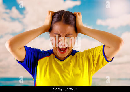 Giovane donna ecuadoriana indossando maratona ufficiale Football Shirt in piedi di fronte alla fotocamera, molto impegnato il linguaggio del corpo la visione di gioco con grande entusiasmo, cielo blu e nuvole sullo sfondo Foto Stock